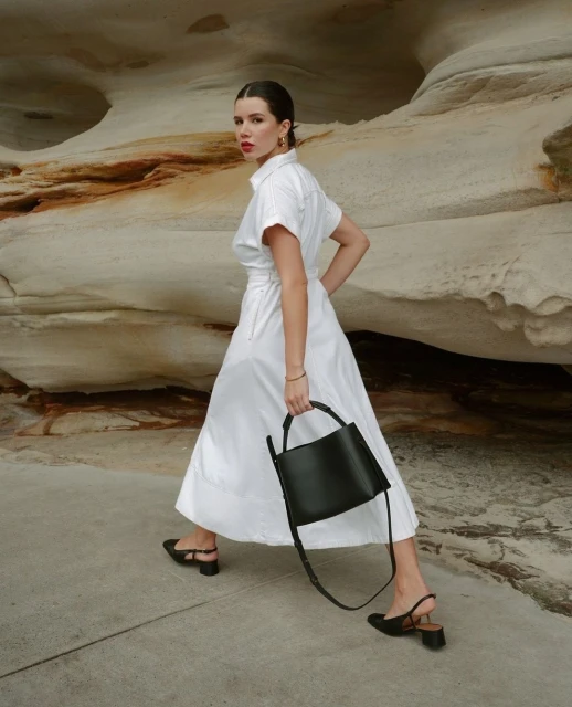 A woman in a white dress walks and carries a black handbag in front of a tan rocky wall on a sunny day, showcasing a fashion-editorial style.