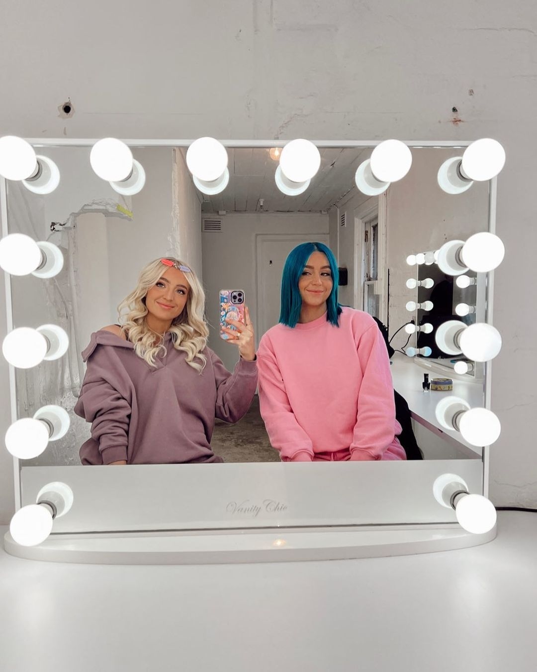 Two smiling young women pose in front of a vanity mirror with illuminated light bulbs in an indoor setting, capturing a vibrant and stylish moment.