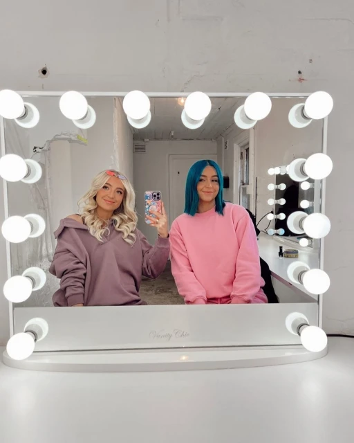 Two smiling young women pose in front of a vanity mirror with illuminated light bulbs in an indoor setting, capturing a vibrant and stylish moment.