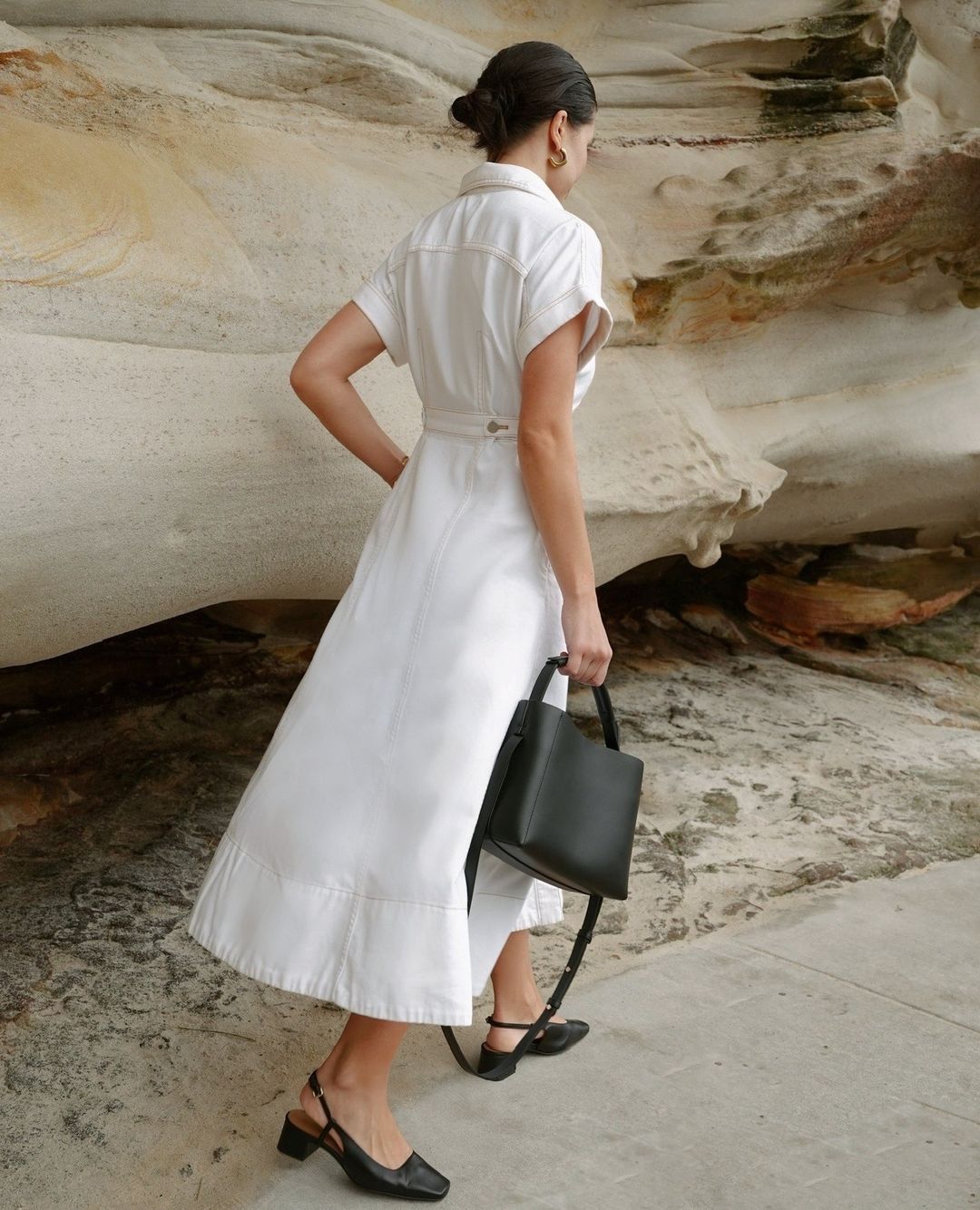 A woman in a white dress and black shoes walks on a stone surface, carrying a black purse with tan and white rock formations behind her.