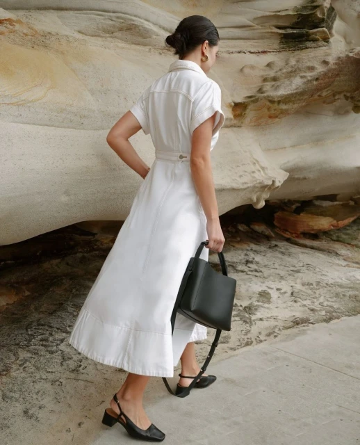 A woman in a white dress and black shoes walks on a stone surface, carrying a black purse with tan and white rock formations behind her.