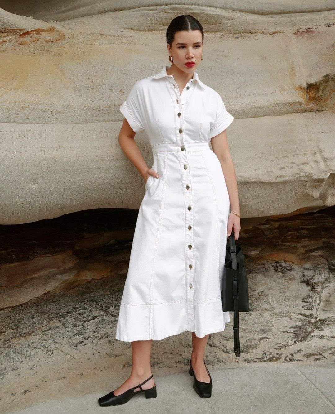 Woman in white collared button-down midi dress with short sleeves, black block heels, and a black tote bag poses against a rock.