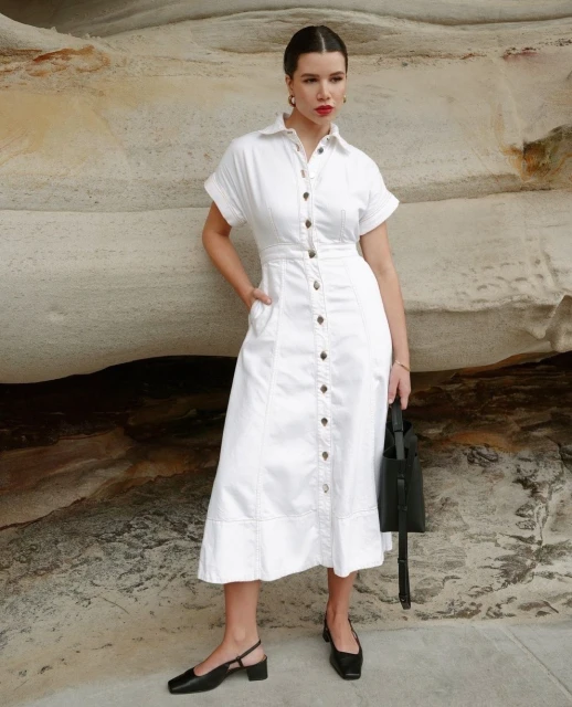 Woman in white collared button-down midi dress with short sleeves, black block heels, and a black tote bag poses against a rock.