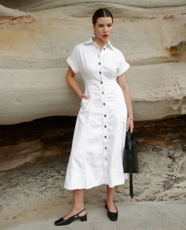Woman in white collared button-down midi dress with short sleeves, black block heels, and a black tote bag poses against a rock.