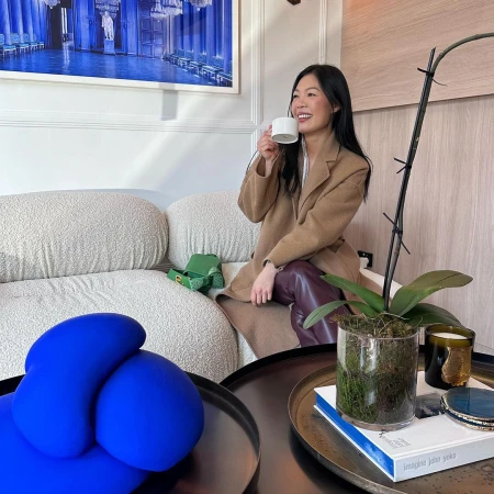 East Asian woman in camel coat holding white coffee cup seated on a white boucle sofa indoors in Sydney