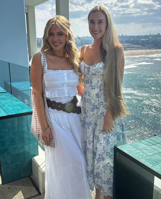 Two women pose in floral and white dresses on a sunny day by the ocean, with city buildings faintly visible in the distance.