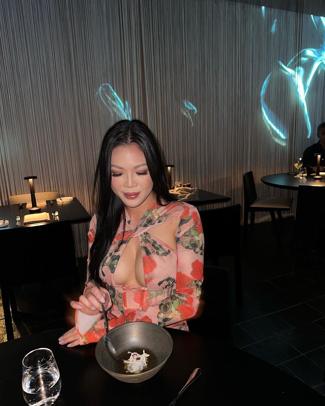 Attractive Asian woman in floral top dining at a restaurant, enjoying soup in a bowl on a black table with a water glass.