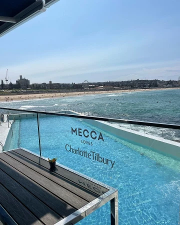 Balcony view overlooking an outdoor coastal swimming pool and sandy beach under a clear blue sky in Sydney.