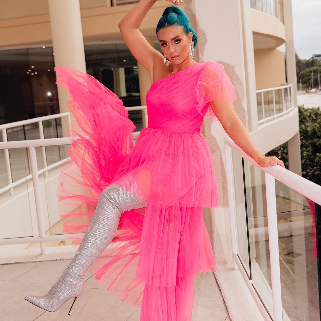 Woman in a bright pink, one-shoulder tulle dress and silver, glittery thigh-high boots poses on a balcony with a building behind her.