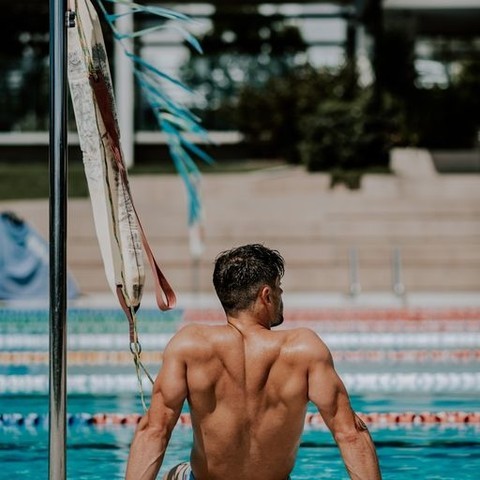 An adult male sits shirtless at the edge of an outdoor swimming pool with visible rippled water and a building backdrop on a sunny day.