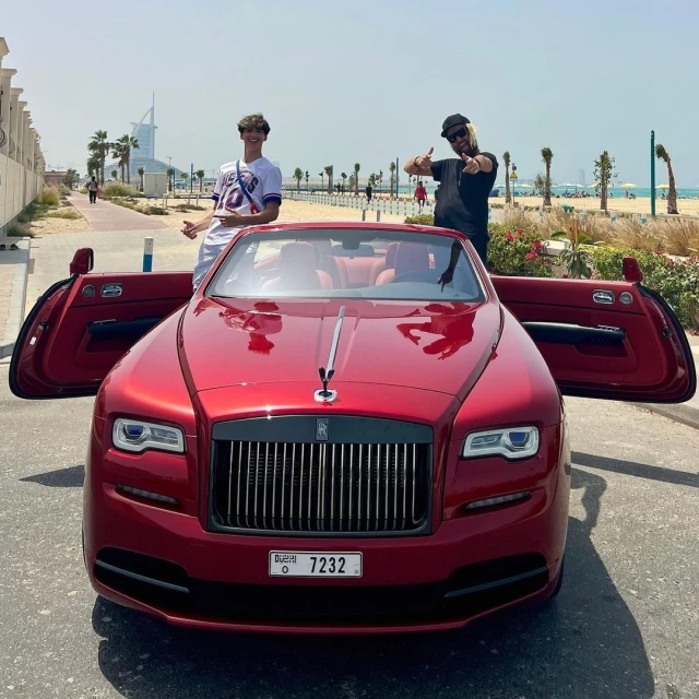 A red Rolls-Royce convertible with two men standing near it on a sunny day, with a beach and Dubai skyline in the background.