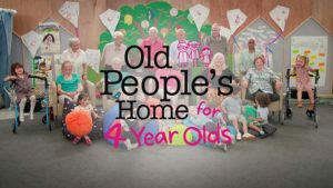A mixed group of elderly people and young children are indoors in a light-filled area, engaging with each other with text overlay.