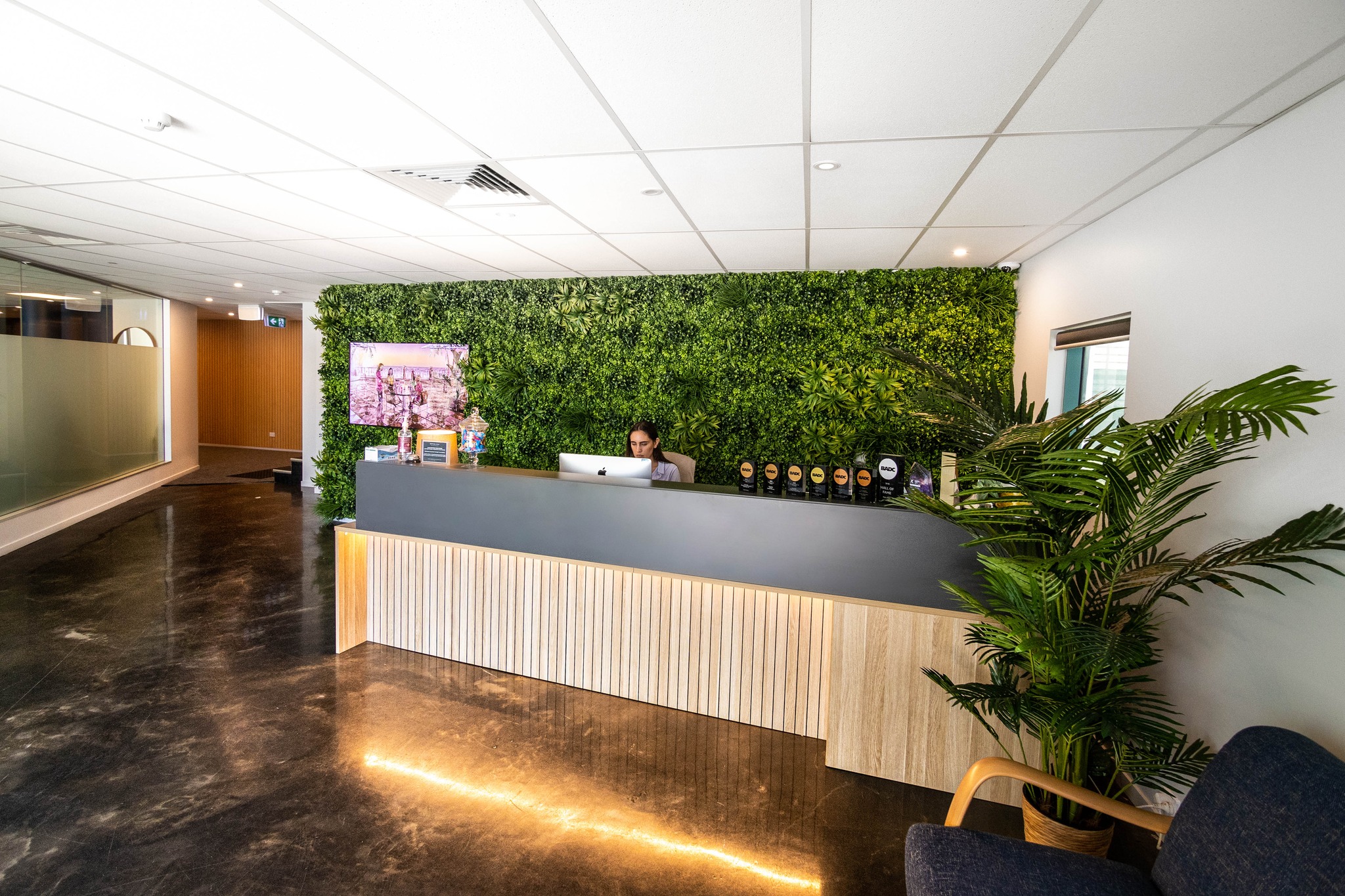 A modern office reception area features a desk with a receptionist, a living plant wall, and a polished floor reflecting light.