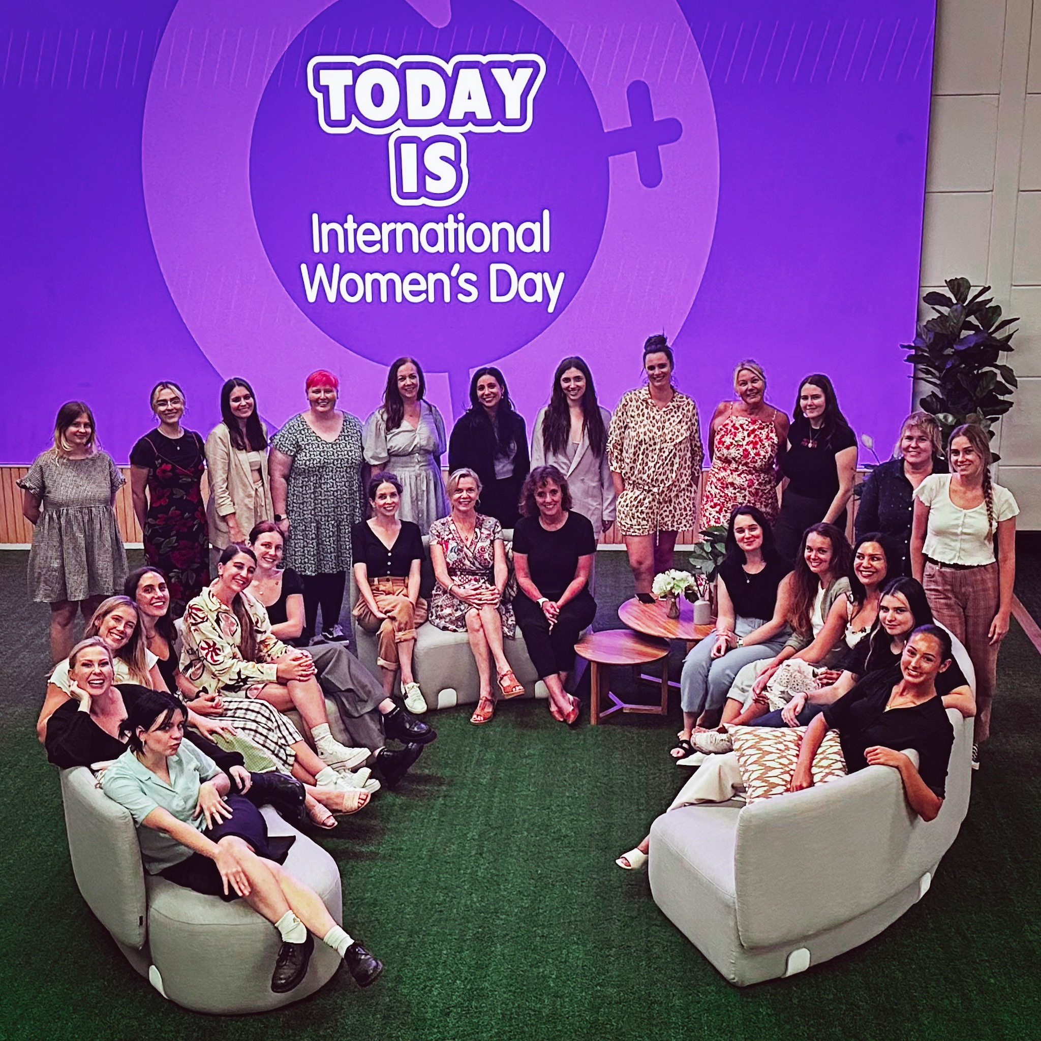 A group of women are posing for a picture in front of a backdrop with the text 'TODAY IS International Women's Day' in a studio.
