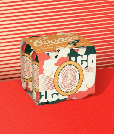 Four-pack of Coopers Regency Park Red Ale cans on a red surface against a red and white striped background in a studio setting.