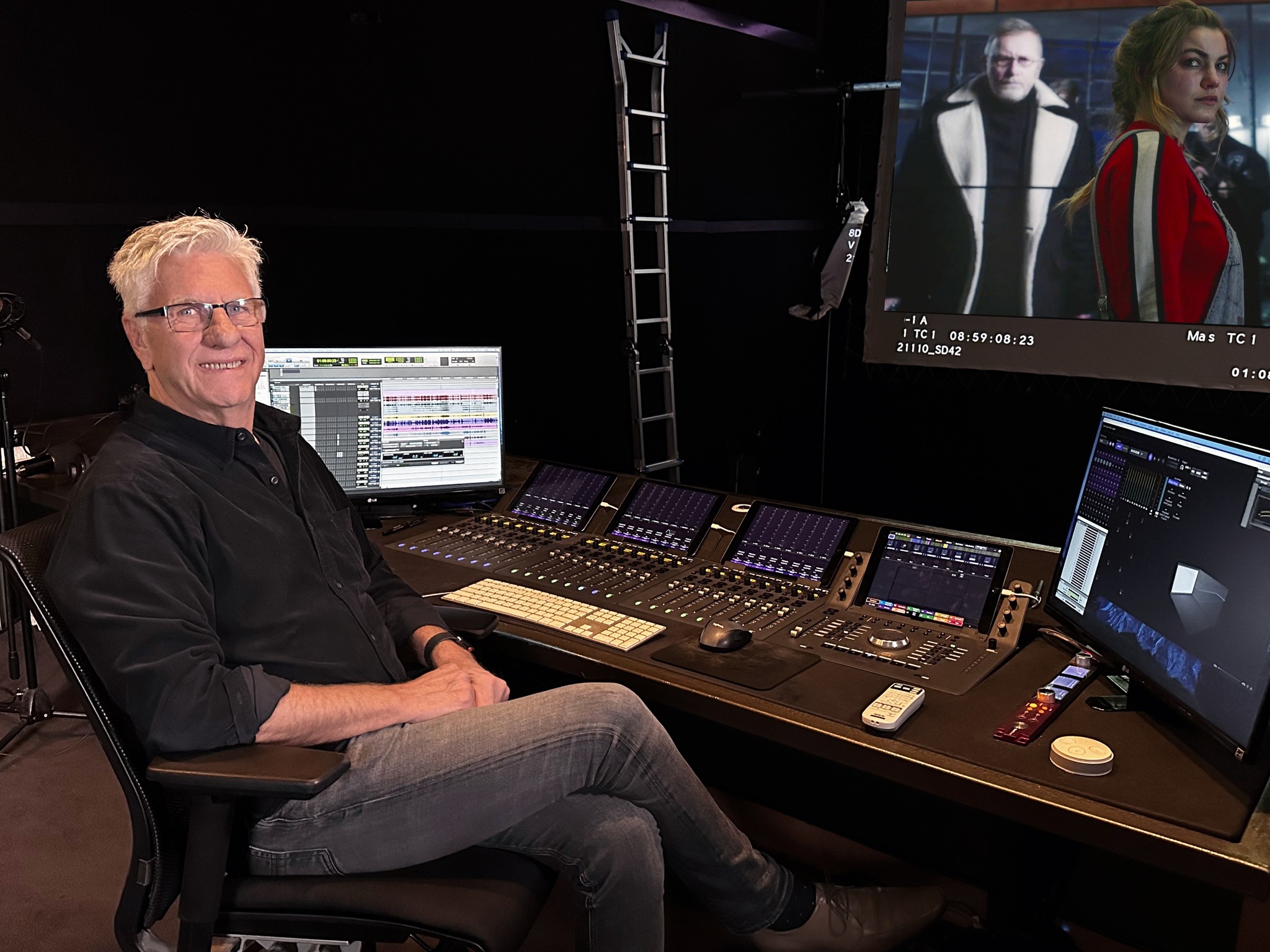 Mature man sits in a dark audio mixing room with digital mixing consoles, computer screens displaying audio tracks, and video playing on a monitor.