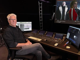A man with white hair seated at a large professional video editing console in a dark studio setting.