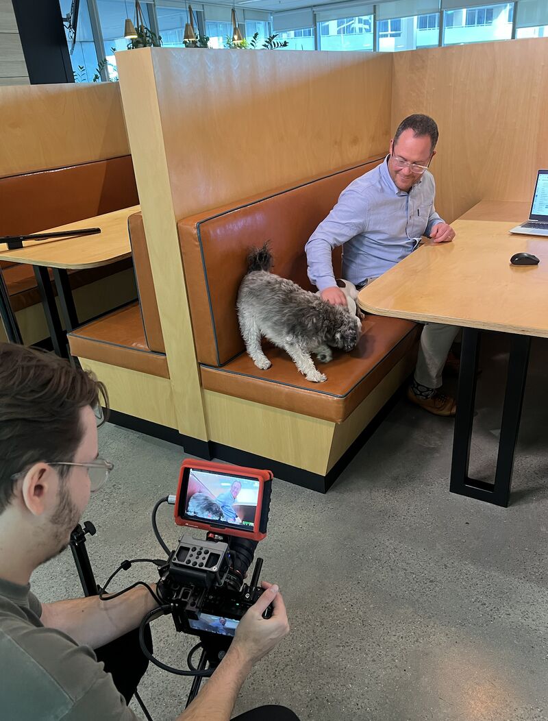 A man pets a dog on a booth seat while being filmed by a videographer in an office. A laptop and coffee cup are on the table.