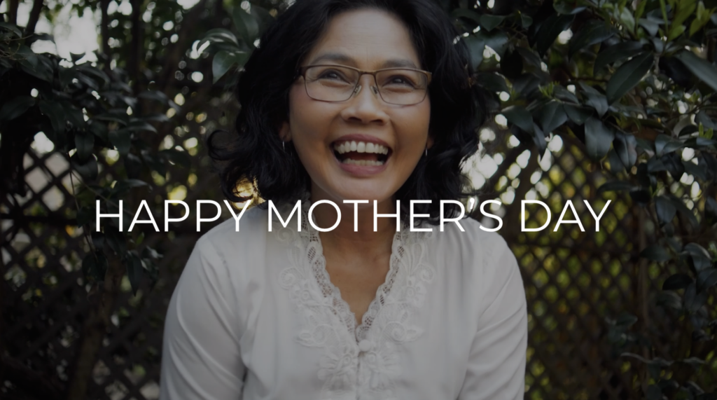 Happy mature woman smiles in a medium shot outdoors, framed by greenery. Wearing glasses and a light top, she exudes joy on Mother's Day.