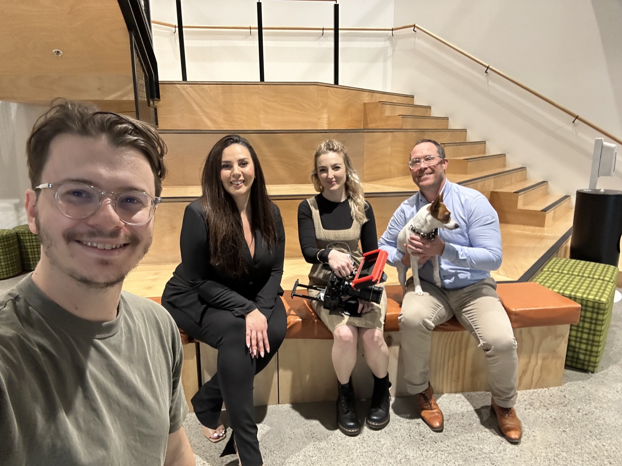 Four adults pose indoors on a bench with a dog. A woman holds a video camera. Stairs, wall and monitor are visible in the background.