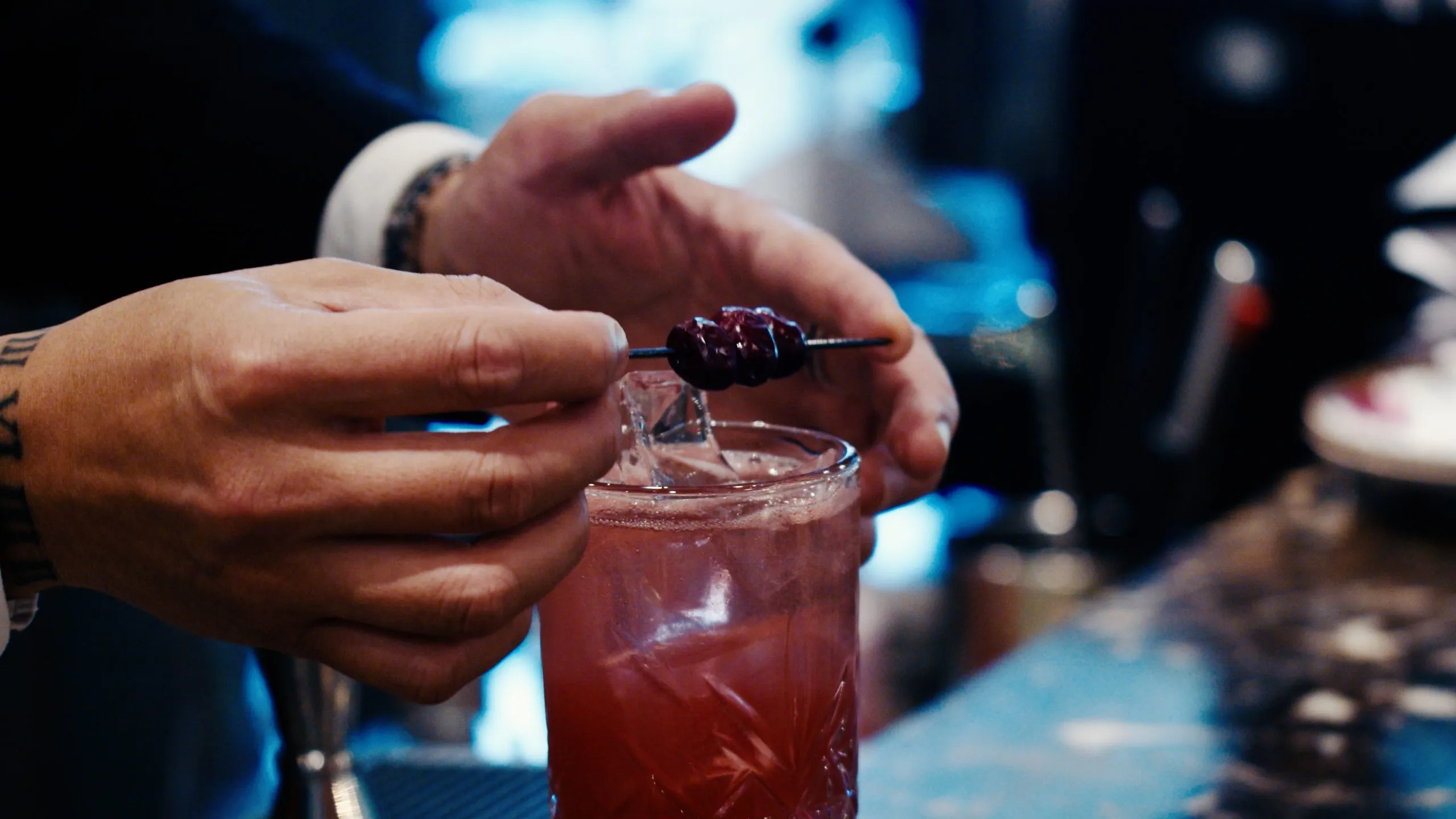 Bartender prepares a red cocktail with ice, garnishing with dark cherries on a skewer, against a blurred dark background.