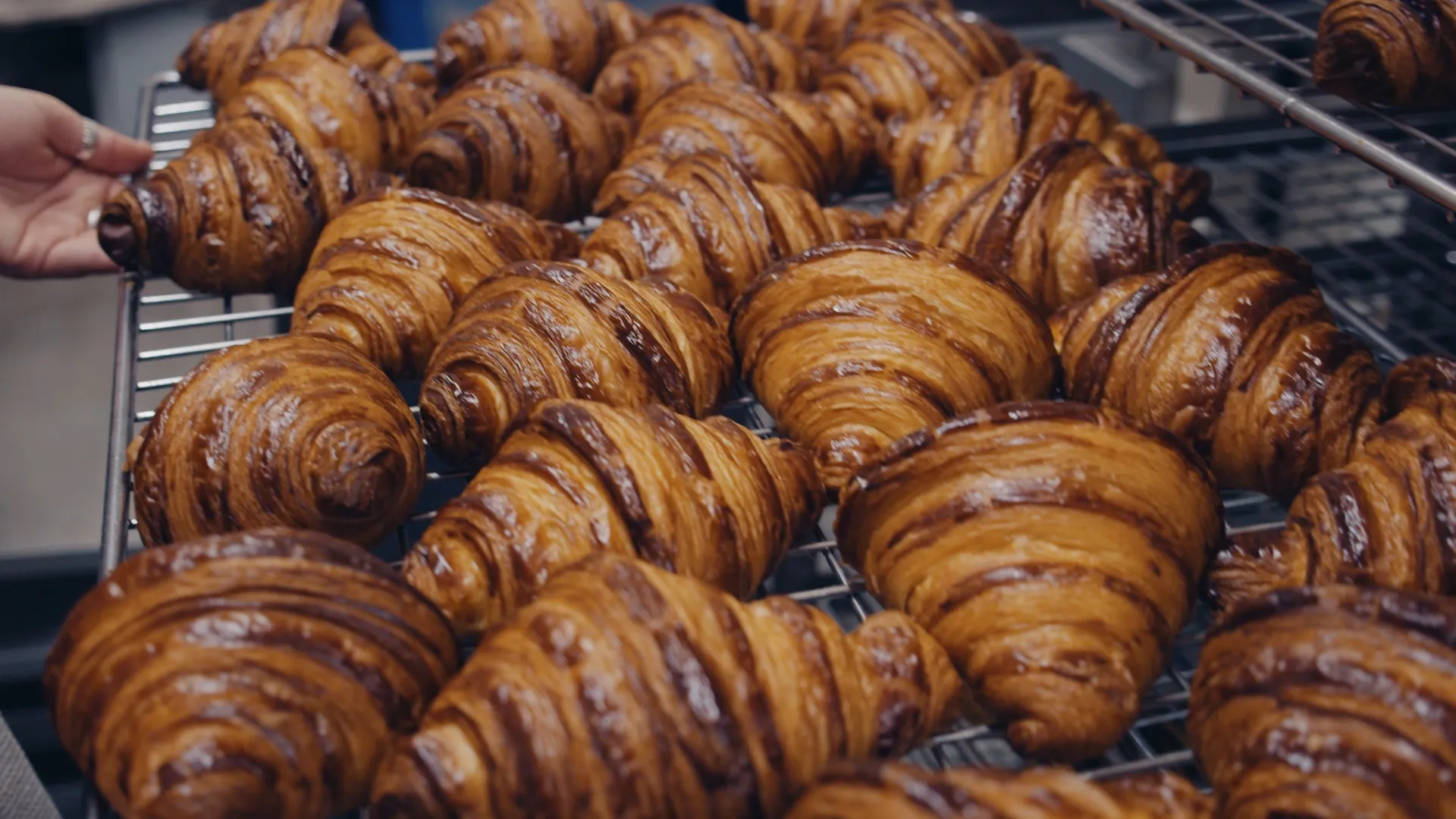 Close-up of an array of freshly baked, golden brown croissants displayed on a metallic cooling rack with hand reaching for one.