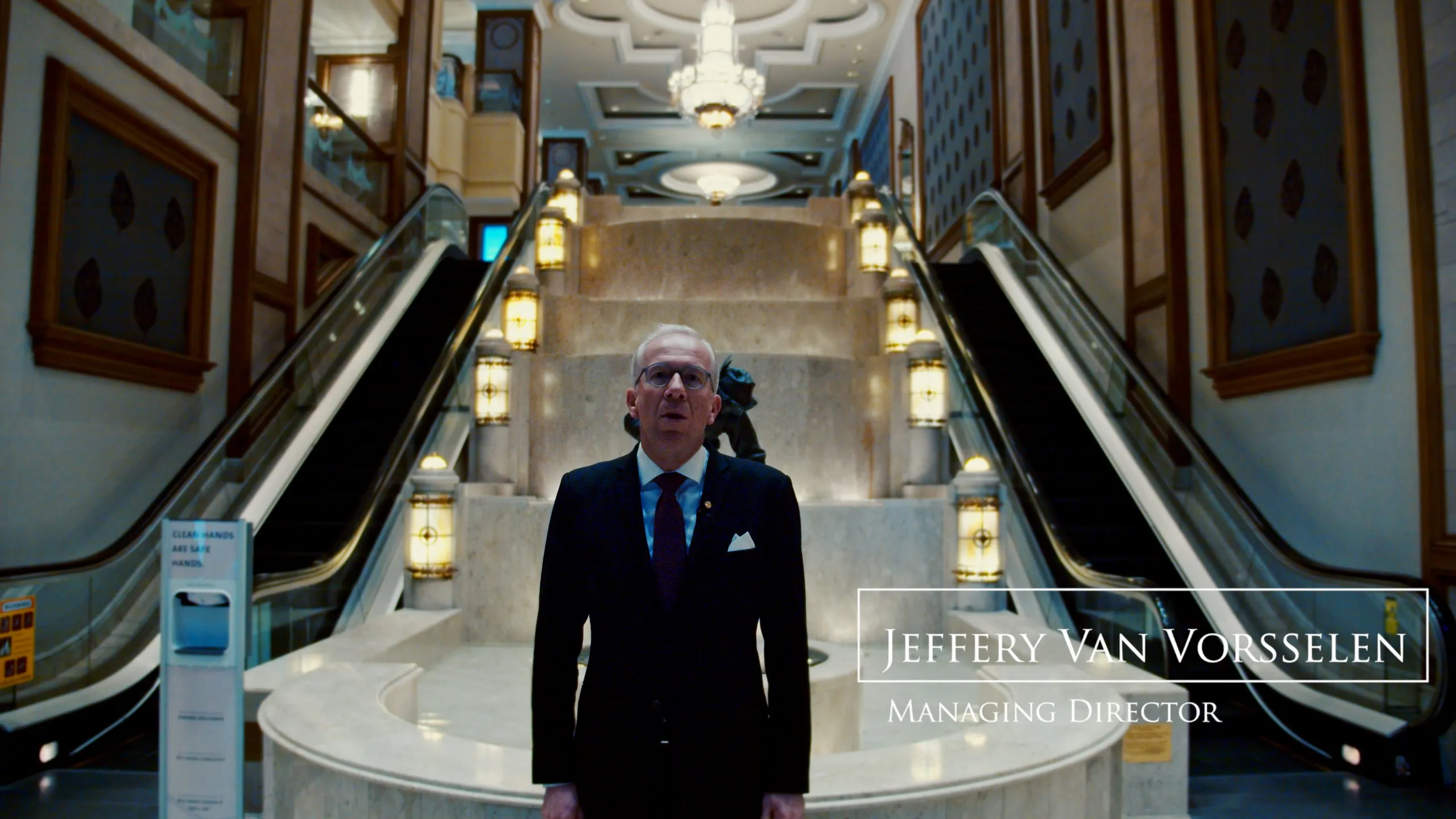 Adult man stands in front of escalators in the opulent lobby of a hotel featuring marble floors, decorative lighting, and ornate architectural details.