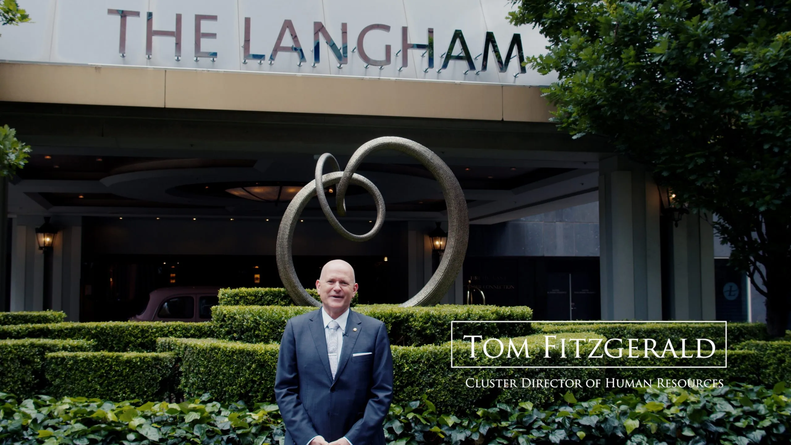 Adult male in a suit stands in front of the Langham Hotel with green shrubs and a looping metal sculpture on a sunny day.