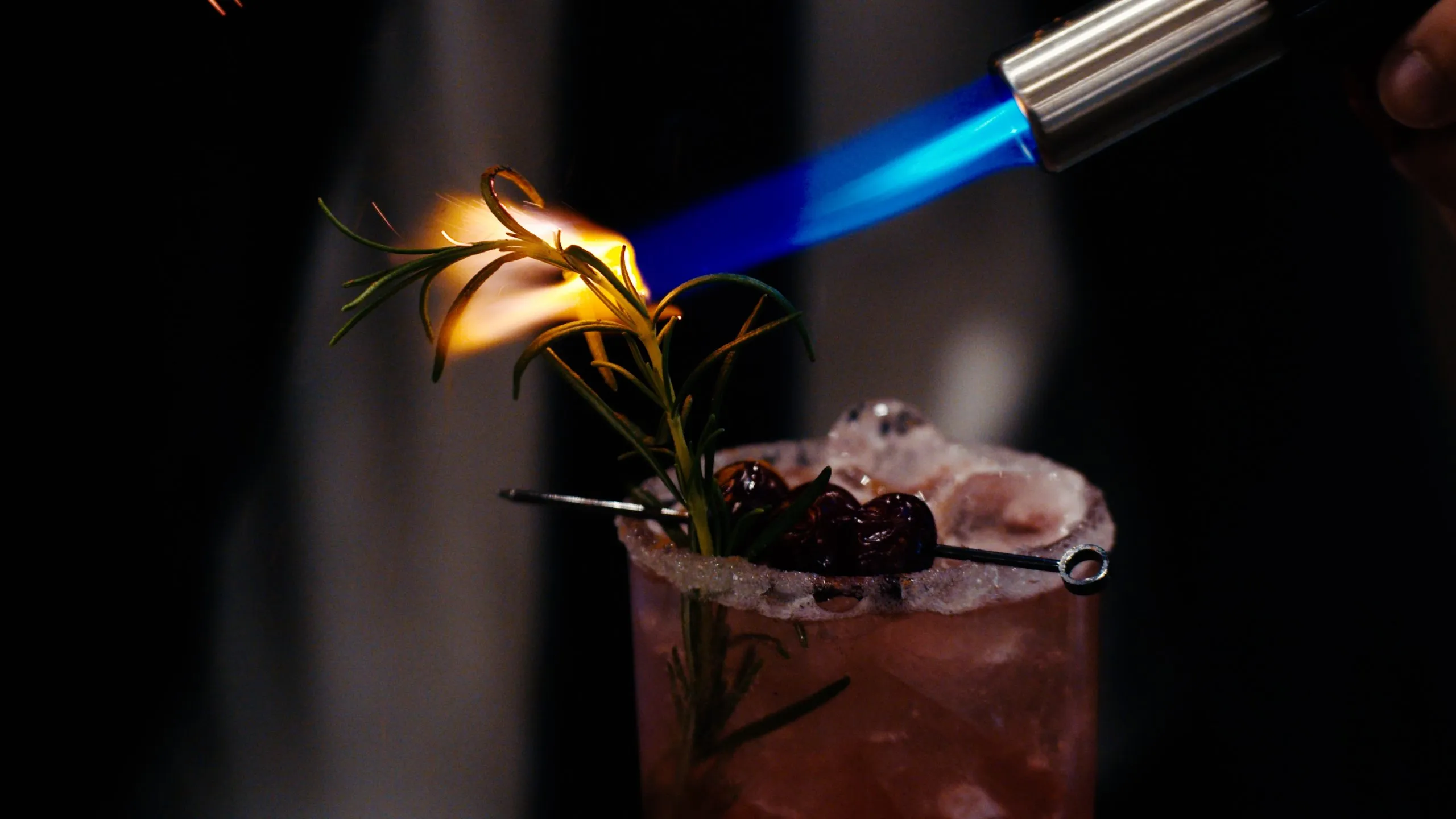 A cocktail is torched with a blue flame in a dark setting, garnished with sugared rim and cherries for a celebratory drink.