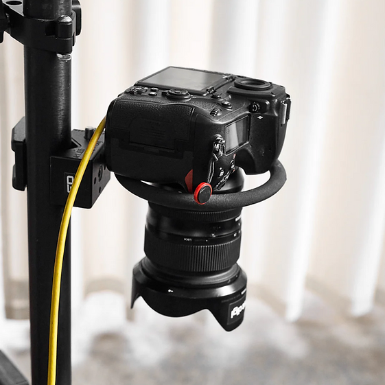 Close-up of a black digital camera attached to a black metal pole, featuring a lens and a yellow cable. The background is soft white.