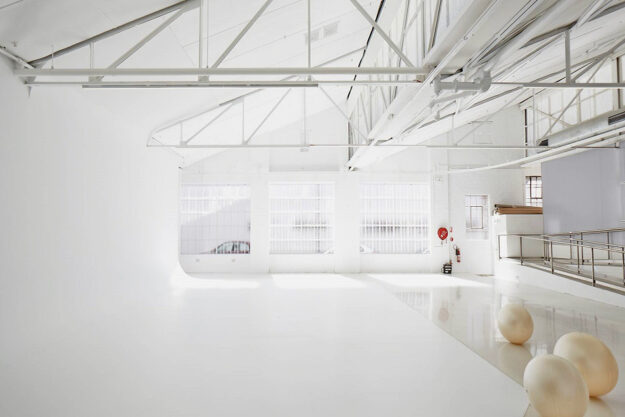 Bright, airy photo studio with white walls, floor, and ceiling. Structural beams visible on the ceiling and windows on the back wall.