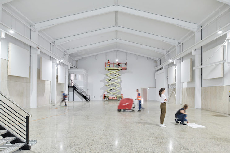Large bright warehouse interior with people working, a scissor lift, concrete floors, and exposed rafters and beams creating geometric patterns.