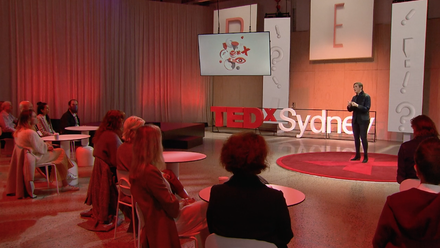 A woman speaks on stage at a TEDx Sydney event with an audience, screen displays, and the TEDx Sydney logo prominent.