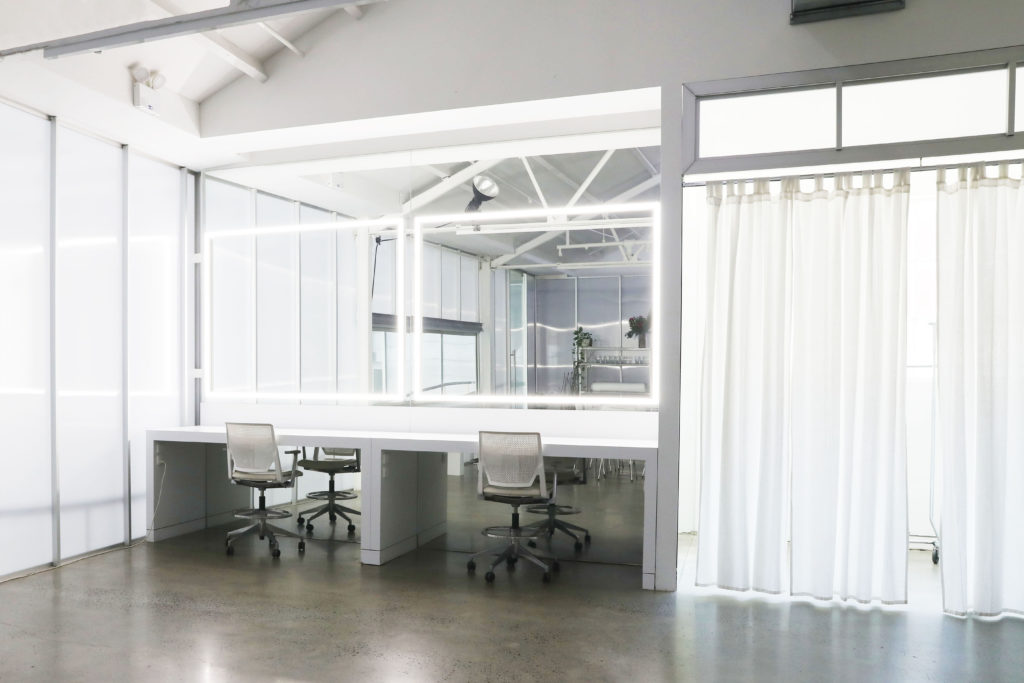Bright photo studio space with white makeup stations, rolling chairs, white curtains, and large mirror area for fashion or photography.