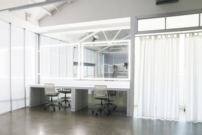 Bright photo studio space with white makeup stations, rolling chairs, white curtains, and large mirror area for fashion or photography.