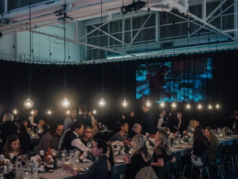 Wide shot of a crowded banquet dinner in an industrial warehouse venue with rows of pendant lights hanging overhead.