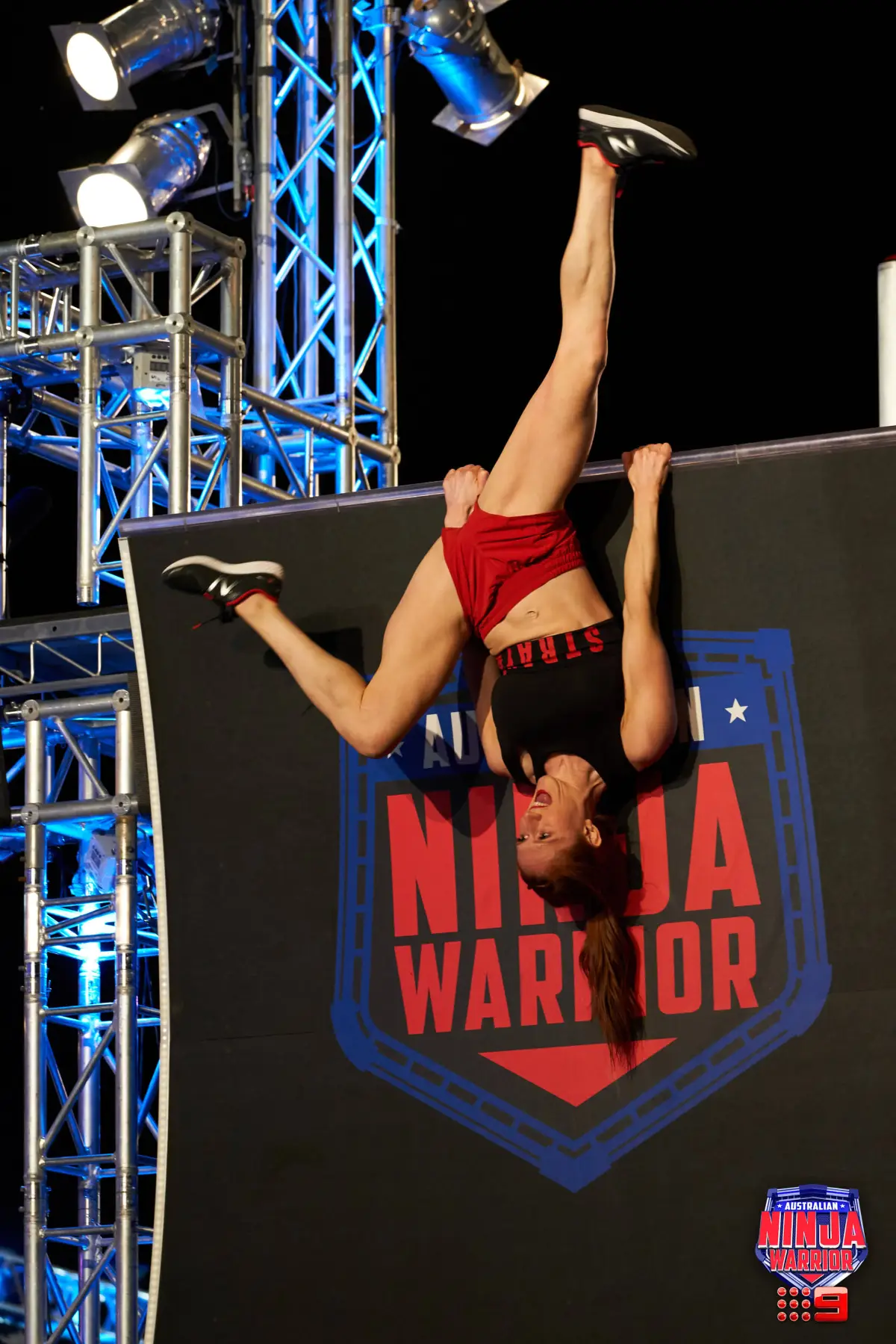 Female athlete competes on an obstacle course, hanging upside down from a bar with her legs extended in a fitness and sports challenge.