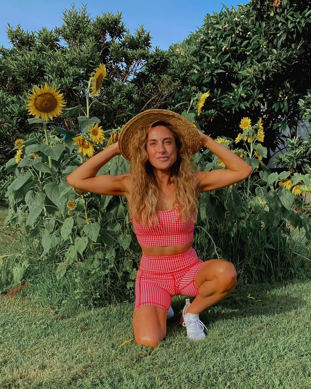 Woman in a red and white gingham outfit kneeling in front of sunflowers in a grassy outdoor setting with a blue sky overhead.