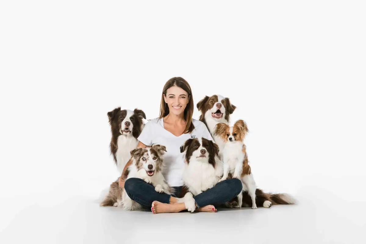 Smiling woman sits barefoot with five dogs of different breeds in a bright studio, exuding warmth and a close bond with her canine companions.