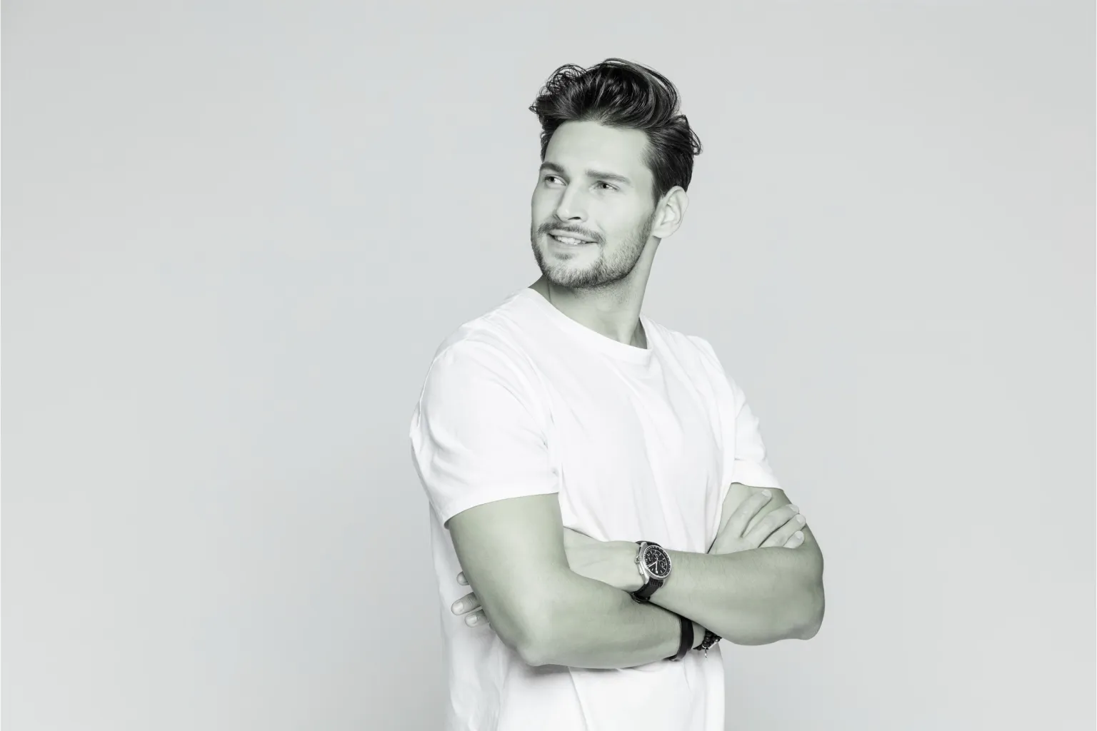 Man with folded arms wearing a white t-shirt and wristwatch looking away against a light grey background.