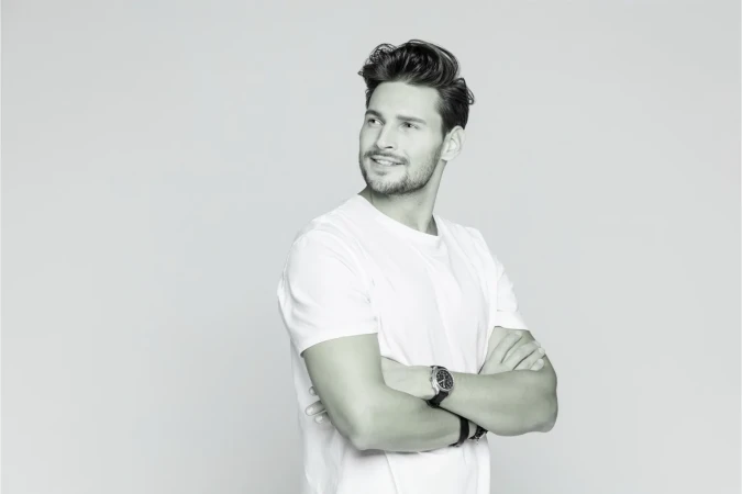 Man with folded arms wearing a white t-shirt and wristwatch looking away against a light grey background.
