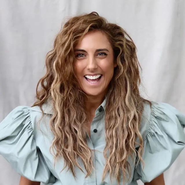 Close-up shot of a woman with curly brown hair smiling broadly while wearing a light green shirt with large, puffed sleeves.