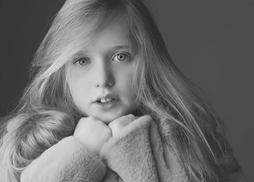 Portrait of a young girl with blonde hair and blue eyes, wearing a light-colored, textured sweater, set against a plain gray backdrop.