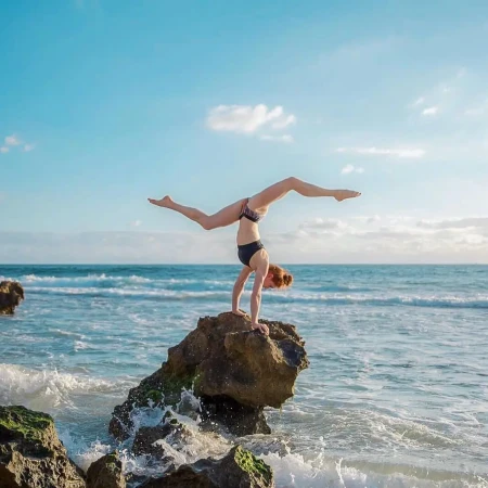 Female athlete performing a handstand on a rock at the beach, showcasing fitness and balance against a backdrop of the ocean and sky.