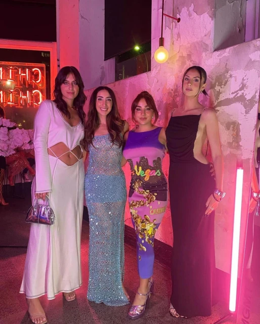Four women in formal dresses at an indoor event. The group stands close together against a textured wall with neon sign and light.