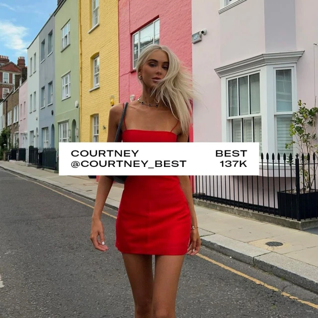 An adult woman with long blonde hair stands on a city street in a red dress in front of colorful row houses on a sunny day.