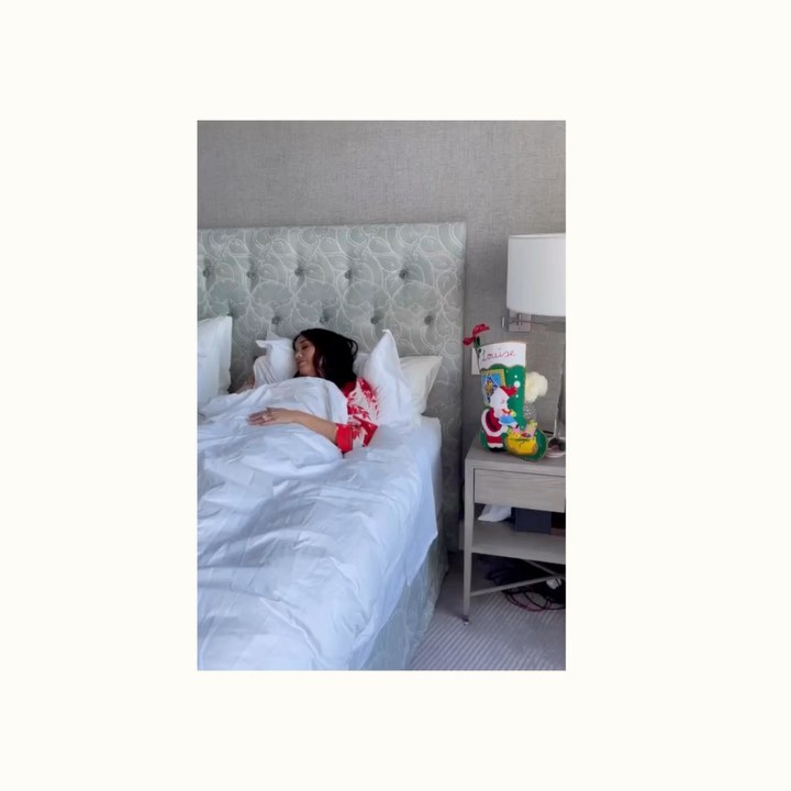 Adult female rests in a bedroom setting, beneath white bed linens. A nightstand with a lamp and Christmas stocking is nearby.