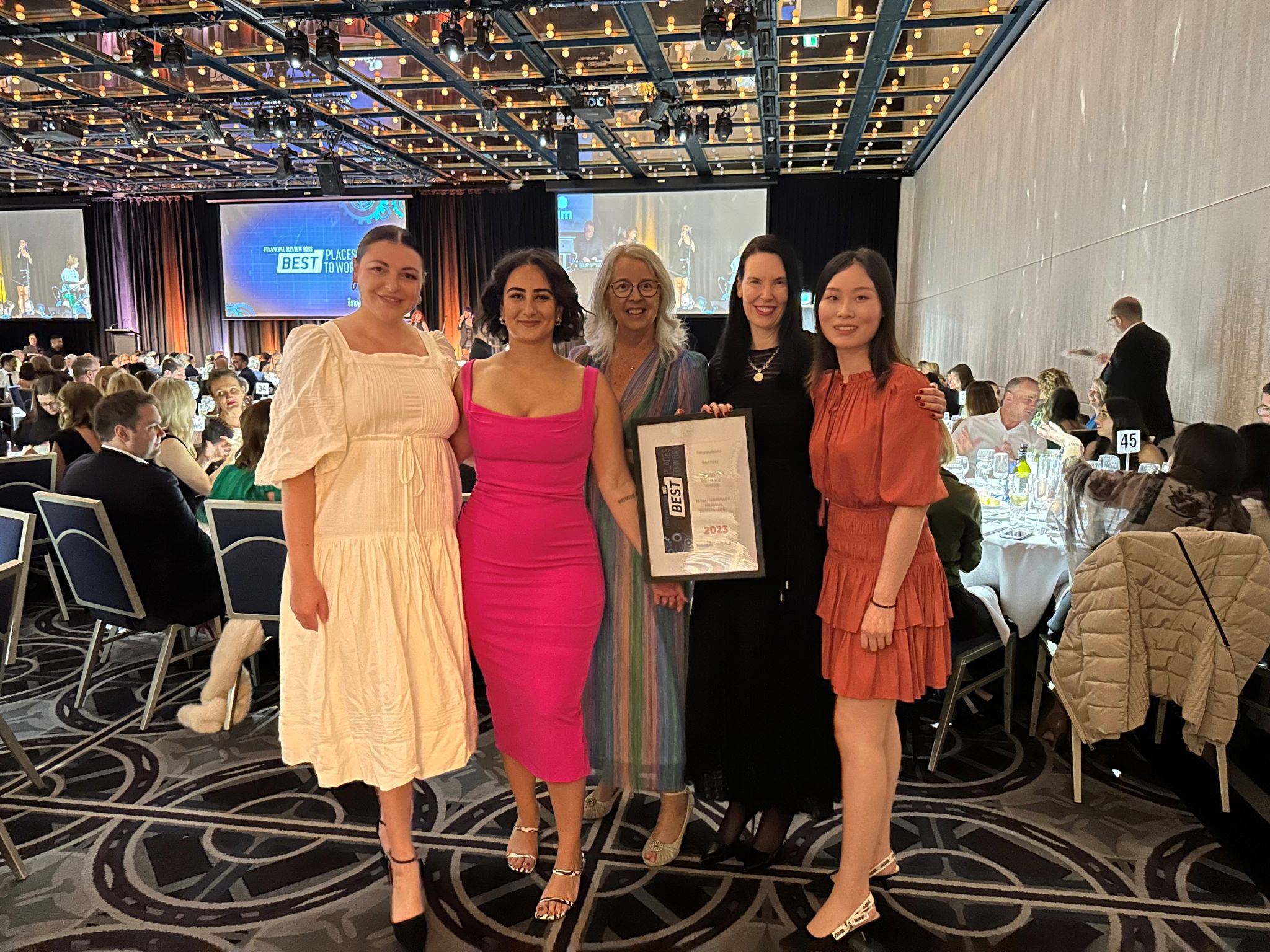 Group of five women posing indoors, holding an award certificate. They are wearing dresses in a well-lit, large room with tables and people.