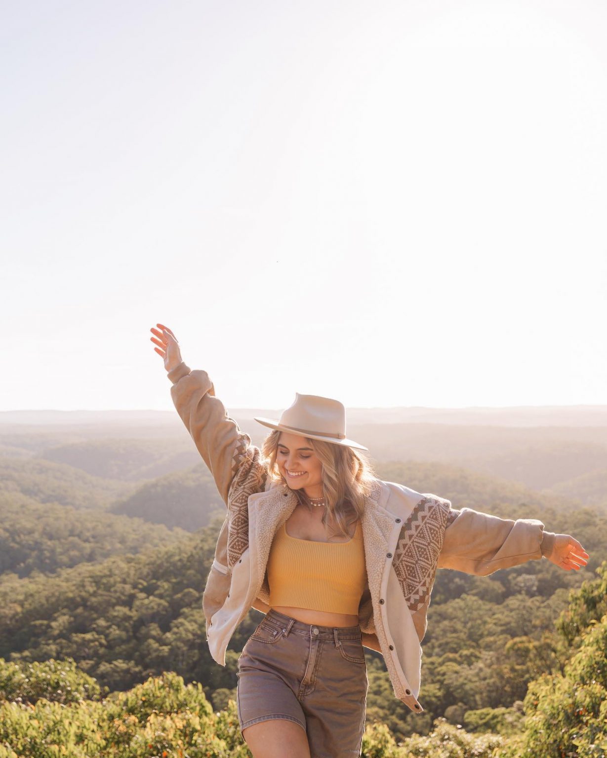 Woman in casual outfit stands with arms outstretched amidst a lush green forest backdrop, bathed in soft sunlight, creating a serene scene.