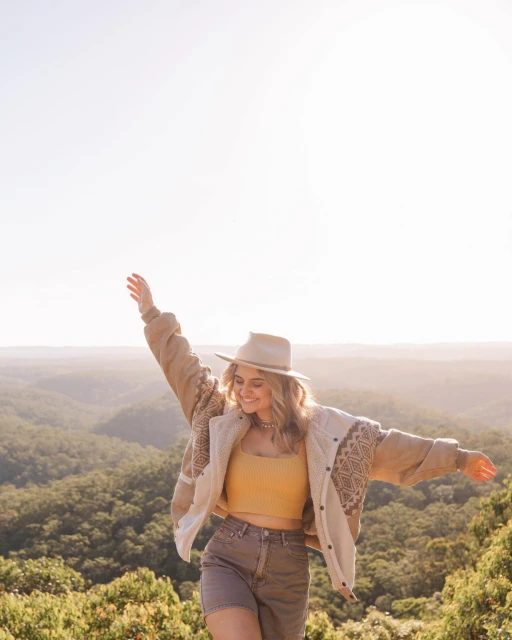 Woman in casual outfit stands with arms outstretched amidst a lush green forest backdrop, bathed in soft sunlight, creating a serene scene.